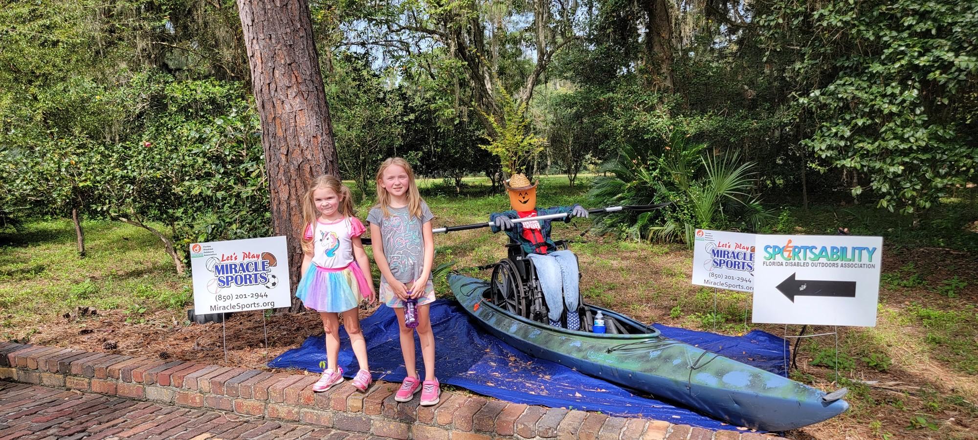 two small children standing in front of a scarecrow in a wheelchair in a kayak
